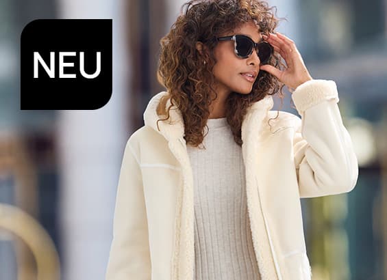 NEU – Frau mit lockigen Haaren trägt hellen Mantel und Sonnenbrille und blickt zur Seite