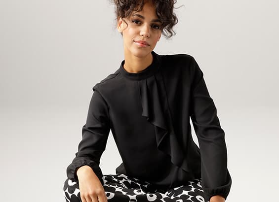 Frau in schwarzer Bluse mit Rüschen, sitzend, trägt gemusterte Hose.