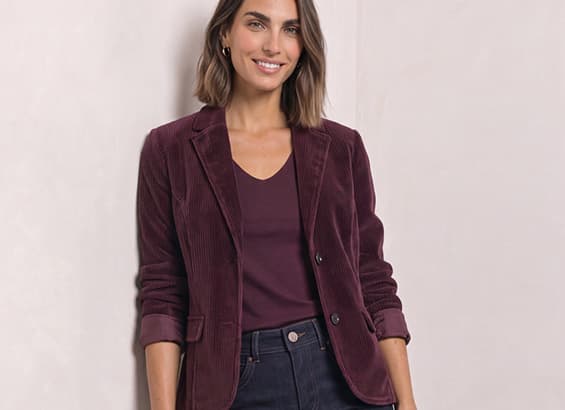 Frau mit schulterlangem Haar trägt bordeauxfarbenen Blazer über passendem Oberteil und dunkler Jeans, steht lächelnd vor hellem Hintergrund